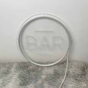 Op de afbeelding: Een wit rond bord met het woord "BAR" gegraveerd in het midden. Het bord is verlicht met witte LED-verlichting en heeft een wit snoer.