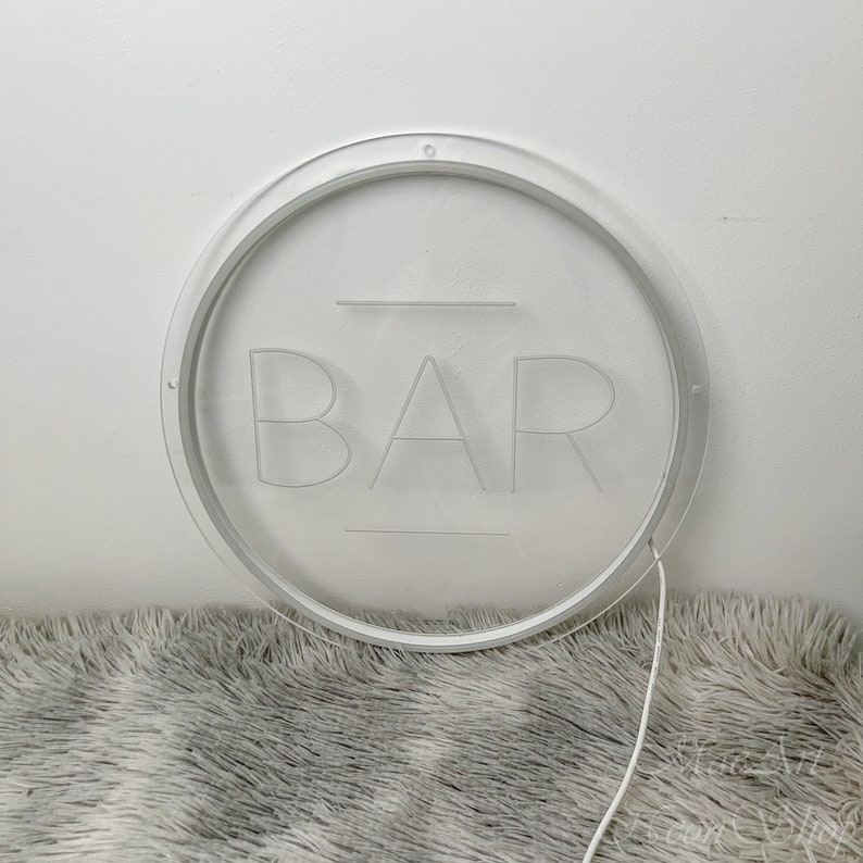 Op de afbeelding: Een wit rond bord met het woord "BAR" gegraveerd in het midden. Het bord is verlicht met witte LED-verlichting en heeft een wit snoer.