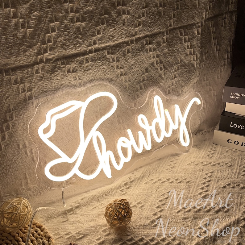 May include: White neon sign with the word "Howdy" in a cursive font. The sign is on a clear acrylic base and is lit up.