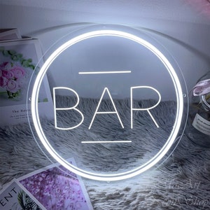 Op de afbeelding: Een wit neon-bord met een ronde lijst en het woord "BAR" in het midden. Het bord ligt op een pluche grijs oppervlak.