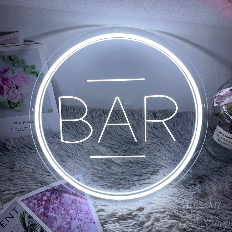 Op de afbeelding: Een wit neon-bord met een ronde lijst en het woord "BAR" in het midden. Het bord ligt op een pluche grijs oppervlak.