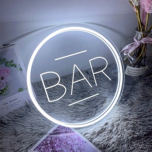 Op de afbeelding: Wit neon-bord met een ronde lijst en het woord "BAR" in witte neon-letters.