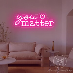 May include: A pink neon sign with the words "you matter" in a cursive font and a heart symbol. The sign is mounted on a white wall above a light pink sofa and a white chair. The sign is from Mac Art Neon Shop.