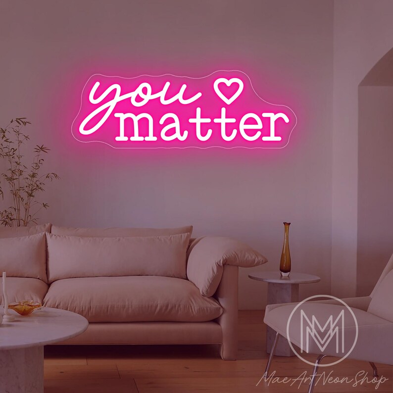 May include: A pink neon sign with the words "you matter" in a cursive font and a heart symbol. The sign is mounted on a white wall above a light pink sofa and a white chair. The sign is from Mac Art Neon Shop.