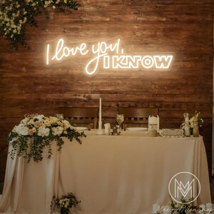 May include: A neon sign displaying the phrase "I love you, I know" in a cursive font, set against a wooden backdrop. The sign is positioned above a wedding reception table, adorned with flowers and candles.