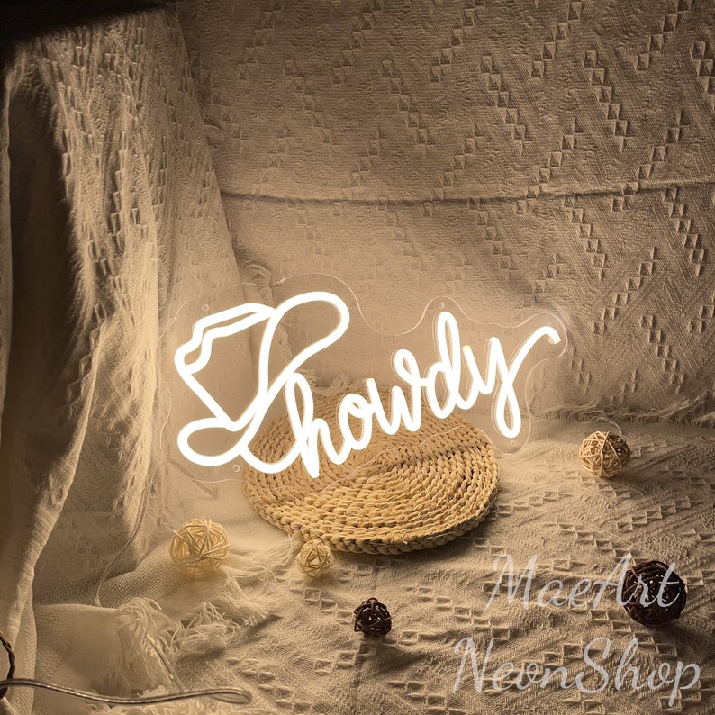 May include: White neon sign with the word "Howdy" in a cursive font, with a cowboy hat design to the left of the word. The sign is sitting on a woven straw mat.
