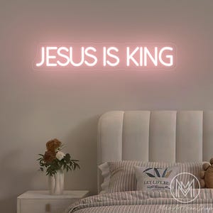 K&ouml;nnte beinhalten: Ein rosafarbenes Neonzeichen mit der Aufschrift &bdquo;JESUS IS KING&ldquo; h&auml;ngt &uuml;ber einem Bett mit einem wei&szlig;en Kopfteil. Links vom Bett befindet sich ein Nachttisch mit einer Blumenvase. Der Raum hat eine weiche, neutrale Farbpalette.