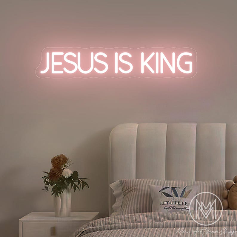 K&ouml;nnte beinhalten: Ein rosafarbenes Neonzeichen mit der Aufschrift &bdquo;JESUS IS KING&ldquo; h&auml;ngt &uuml;ber einem Bett mit einem wei&szlig;en Kopfteil. Links vom Bett befindet sich ein Nachttisch mit einer Blumenvase. Der Raum hat eine weiche, neutrale Farbpalette.
