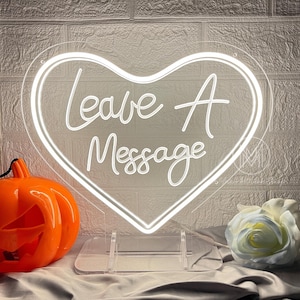 Puede incluir: Un letrero de neón en forma de corazón con las palabras "Leave A Message" en fuente cursiva. Una calabaza naranja con una cara tallada y una rosa blanca están a cada lado del letrero.