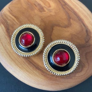 May include: A pair of vintage clip-on earrings. Each earring features a round, gold-toned frame with a black center and a vibrant red, circular stone. The earrings have a textured, ridged border and a classic, elegant design.