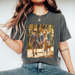 May include: Dark gray t-shirt with a family photo print. The photo shows a family walking in a forest. The shirt is worn with blue jeans and white sneakers. A gold necklace and bracelet are also visible.