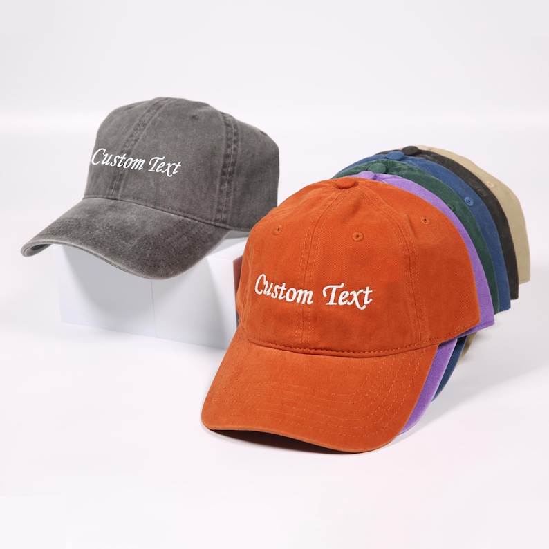 May include: A collection of baseball caps in various colors, including orange, gray, and navy. Each cap features the embroidered text "Custom Text" in white. The caps are arranged in a row, showcasing their different hues.