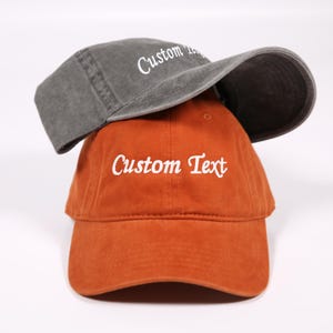 May include: Two baseball caps, one gray and one orange, are stacked. The gray cap is on top, with the text "Custom" in white. The orange cap below has "Custom Text" in white. Both caps have a curved brim.