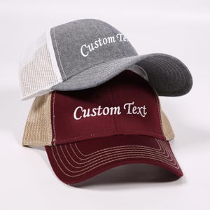 May include: Two baseball caps, one gray and one burgundy, stacked on top of each other. The gray cap has a white mesh back and the burgundy cap has a tan mesh back. Both caps have the words "Custom Text" embroidered in white.