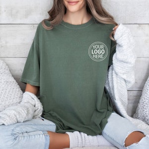 May include: A woman wearing a green oversized t-shirt with a white circle on the left chest that says "YOUR LOGO HERE". She is sitting on a white couch with a white blanket.