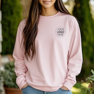 May include: A light pink long-sleeve shirt with a crew neck. The shirt features a small, circular logo that reads "YOUR LOGO HERE" in black. The sleeves are long, and the shirt appears to be made of a soft material.