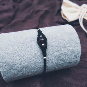 Puede incluir: Un brazalete de cord&oacute;n negro con una cuenta de piedra preciosa de color p&uacute;rpura oscuro. El brazalete est&aacute; sobre una superficie de tela gris.