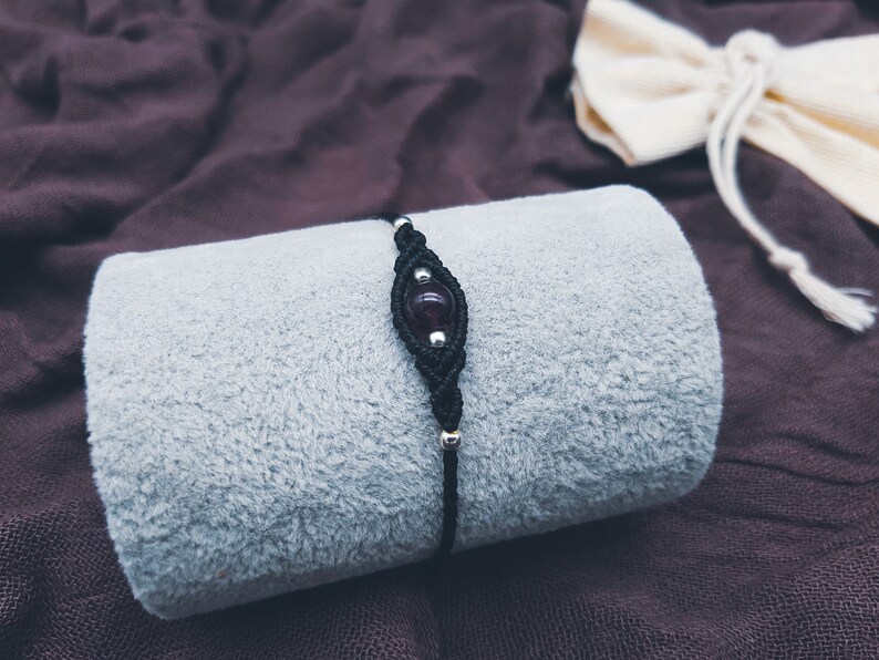 Puede incluir: Un brazalete de cord&oacute;n negro con una cuenta de piedra preciosa de color p&uacute;rpura oscuro. El brazalete est&aacute; sobre una superficie de tela gris.