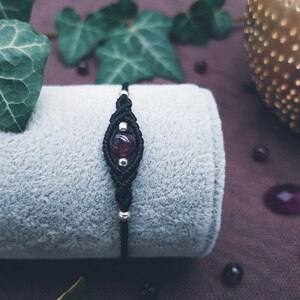 Puede incluir: Un brazalete de macram&eacute; negro con una cuenta de piedra preciosa de color p&uacute;rpura oscuro. El brazalete est&aacute; sobre una superficie de tela gris.