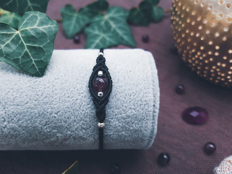Puede incluir: Un brazalete de macram&eacute; negro con una cuenta de piedra preciosa de color p&uacute;rpura oscuro. El brazalete est&aacute; sobre una superficie de tela gris.