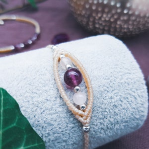 Puede incluir: Un brazalete delicado con un cordón de color crema tejido con pequeñas cuentas plateadas y tres cuentas de piedras preciosas: una amatista púrpura, un cuarzo rosa y otra amatista púrpura.