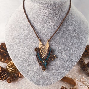 Puede incluir: Un collar de cordón marrón con un colgante dorado y turquesa. El colgante está hecho de un diseño geométrico con un cordón tejido y cuentas.
