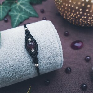 Puede incluir: Un brazalete de macram&eacute; negro con una cuenta de piedra preciosa de color p&uacute;rpura oscuro. El brazalete est&aacute; sobre una superficie de tela gris.