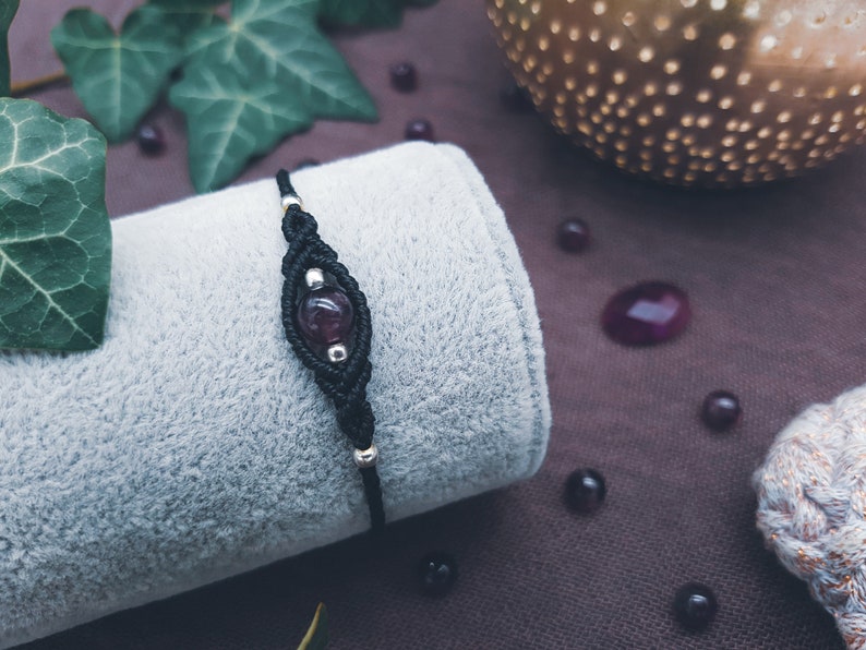 Puede incluir: Un brazalete de macram&eacute; negro con una cuenta de piedra preciosa de color p&uacute;rpura oscuro. El brazalete est&aacute; sobre una superficie de tela gris.