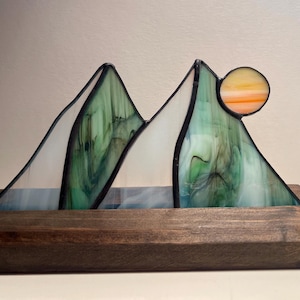 May include: Stained glass mountain scene with a setting sun. The peaks are green and white, set in a dark wooden base. The sun is a circle of orange and yellow glass. A decorative piece for home decor.