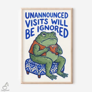 May include: A framed art print showing a grumpy green frog in an orange vest, holding a mug. The frog is seated on a blue patterned ottoman. The text "UNANNOUNCED VISITS WILL BE IGNORED" is written in blue.