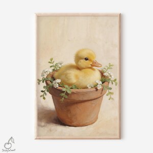 May include: A framed print featuring a fluffy yellow duckling nestled in a terracotta pot filled with white flowers and green foliage. The duckling has an orange beak and is set against a soft, neutral background. The artwork has a vintage, painterly style.