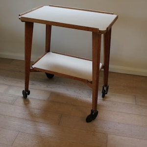 May include: A two-tiered rolling trolley featuring a wooden frame with four black castors. The trolley has a rectangular top surface and a lower shelf, both finished in a smooth, off-white material with raised wooden edges on the top tier.