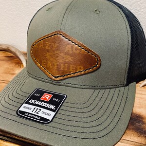 May include: A green and black trucker hat with a brown leather patch. The hat has a black mesh back and a black stitched brim. The hat has a label that reads "EST. 1970 RICHARDSON ORIGINAL 112 TRUCKER ADJUSTABLE".