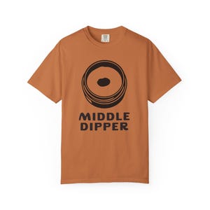 May include: A rust-colored t-shirt featuring a black graphic of concentric circles with a dark center. Below the graphic, the words "MIDDLE DIPPER" are printed in bold, black letters. The shirt has short sleeves and a crew neck.