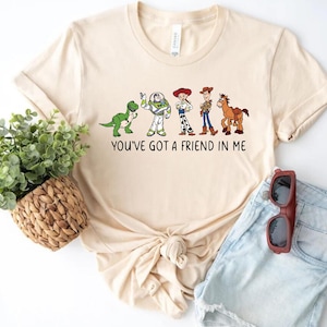 May include: A beige t-shirt with a graphic of the Toy Story characters Rex, Buzz Lightyear, Woody, Jessie, and Bullseye. The text "You've got a friend in me" is printed below the graphic.