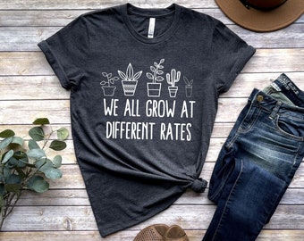 May include: A dark gray t-shirt with a white graphic of potted plants and the text "We all grow at different rates".