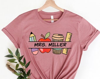 May include: A mauve t-shirt with a graphic design of a pencil, apple, coffee cup, and ruler. The text "MRS. MILLER" is printed on a banner above the graphic.