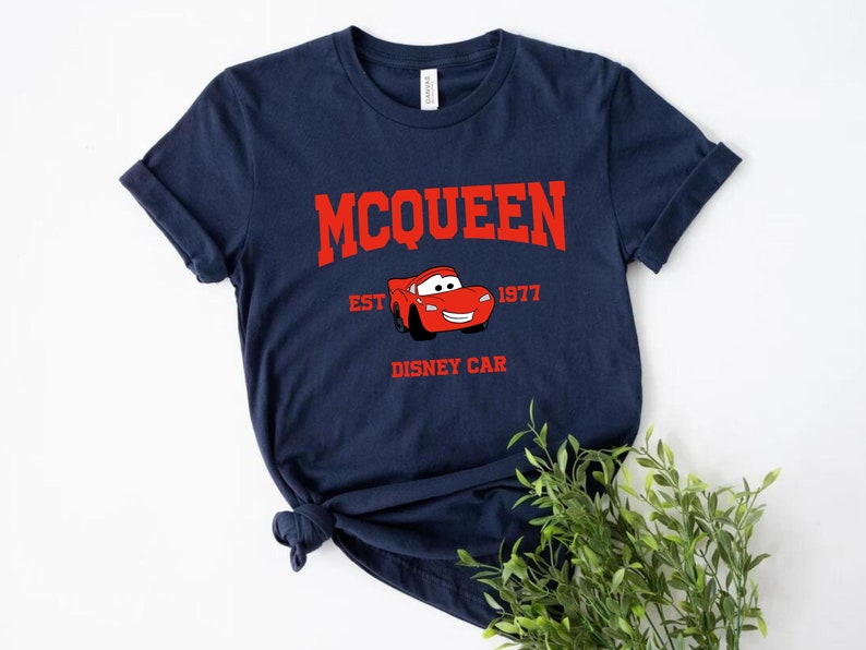 May include: A navy blue t-shirt featuring a red graphic of Lightning McQueen from the Disney film Cars. The text "MCQUEEN EST 1977 DISNEY CAR" is printed on the shirt.