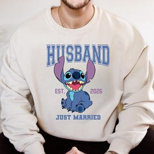 May include: Cream-colored sweatshirt featuring a blue cartoon character with large ears and a wide smile. The word "HUSBAND" is printed above the character in blue block letters, with "EST. 2026" and "JUST MARRIED" below.