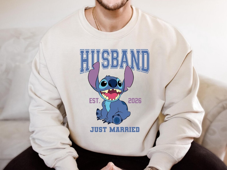 May include: Cream-colored sweatshirt featuring a blue cartoon character with large ears and a wide smile. The word "HUSBAND" is printed above the character in blue block letters, with "EST. 2026" and "JUST MARRIED" below.