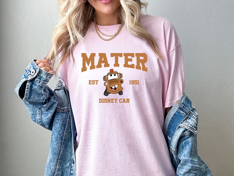 May include: Pink t-shirt featuring a brown graphic of Mater from the Disney film Cars. The graphic includes the text "Mater", "Est. 1951", and "Disney Car".