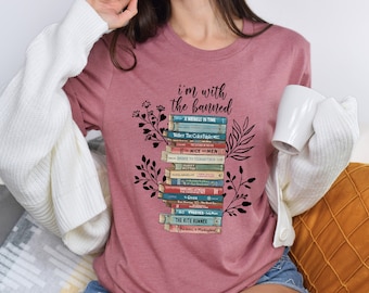 May include: A pink t-shirt with the text "i'm with the banned" and a stack of books with titles like "A Wrinkle in Time", "To Kill a Mockingbird", and "The Kite Runner".