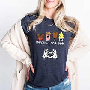 May include: A dark heather grey t-shirt with a graphic of Mickey Mouse ears, popcorn, a Mickey Mouse ice cream bar, and a Dole Whip. The text "Snacking for Two" is printed below the graphic. Mickey Mouse hands are making a heart shape below the text.