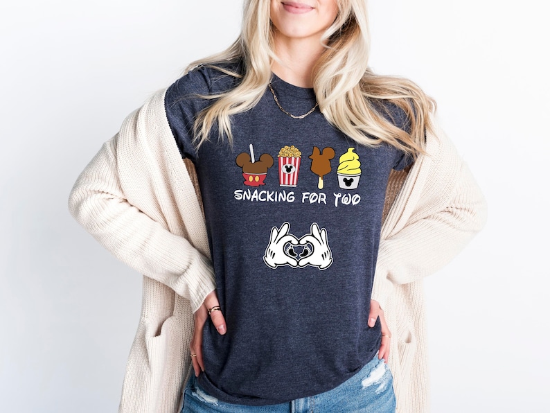 May include: A dark heather grey t-shirt with a graphic of Mickey Mouse ears, popcorn, a Mickey Mouse ice cream bar, and a Dole Whip. The text "Snacking for Two" is printed below the graphic. Mickey Mouse hands are making a heart shape below the text.
