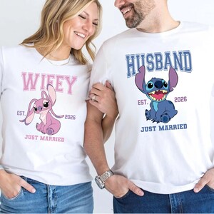 May include: White t-shirts with matching graphics. One shirt says "Wifey" with a pink cartoon character and the other says "Husband" with a blue cartoon character. Both shirts have "EST. 2026" and "JUST MARRIED".