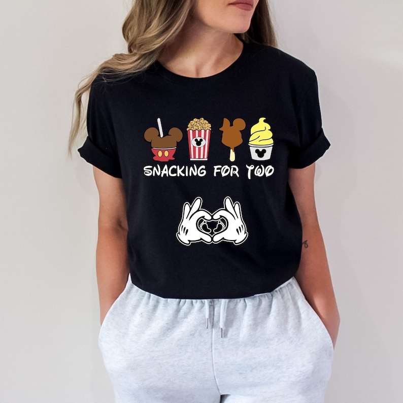 May include: Black t-shirt with a graphic of Mickey Mouse ears, popcorn, a Mickey Mouse ice cream bar, and a Mickey Mouse ice cream cone. The text "Snacking for Two" is below the graphic. Two Mickey Mouse hands are making a heart shape with a baby inside.