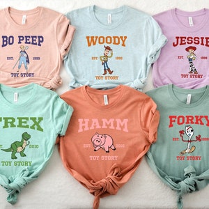 May include: Six different colored t-shirts with Toy Story characters printed on them. The shirts are all folded and laid out on a white surface. The characters include Woody, Jessie, Bo Peep, Rex, Hamm, and Forky. Each shirt has the character's name and the text "Toy Story" printed on it.