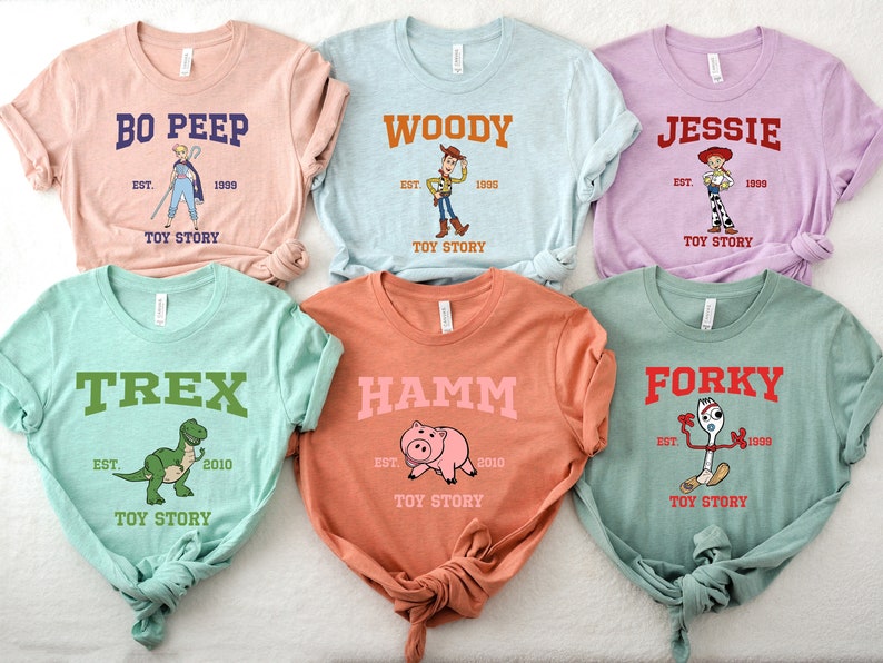 May include: Six different colored t-shirts with Toy Story characters printed on them. The shirts are all folded and laid out on a white surface. The characters include Woody, Jessie, Bo Peep, Rex, Hamm, and Forky. Each shirt has the character's name and the text "Toy Story" printed on it.