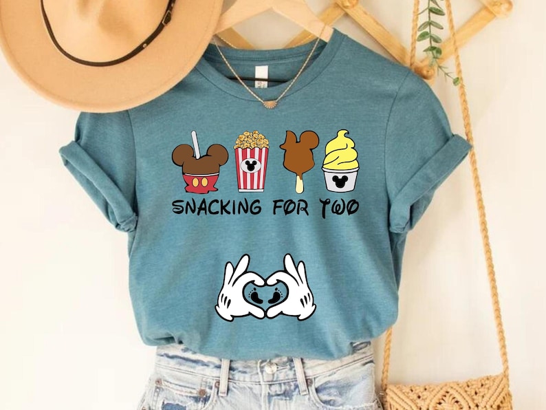 May include: A teal blue t-shirt with a graphic of Mickey Mouse snacks, including popcorn, a Mickey Mouse ice cream bar, and a Mickey Mouse ice cream cone. The text "Snacking for Two" is printed below the snacks.  A pair of Mickey Mouse hands are holding a heart with two baby footprints inside.