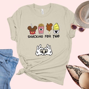May include: A light beige t-shirt with a graphic of Mickey Mouse ears, popcorn, a Mickey Mouse ice cream bar, and a Mickey Mouse ice cream cone. The text "Snacking for Two" is printed below the graphic. Two white Mickey Mouse hands are making a heart shape with their fingers below the text.